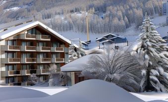 a snow - covered mountain with a ski resort , including several buildings and trees , in the background at Hotel Ambassador Zermatt