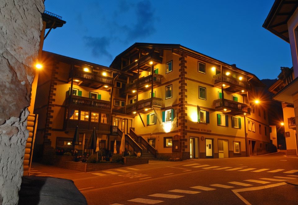 a large , multi - story building with balconies and windows is lit up at night , with the entrance to a parking lot visible in at Hotel Alpino