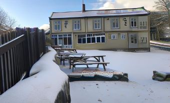 a snowy scene with a large building and several picnic tables in the foreground , all covered with snow at Anglers Arms