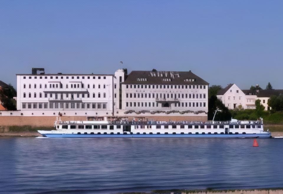 a large white building on the banks of a river , with multiple boats docked along its length at Hotel am Rhein