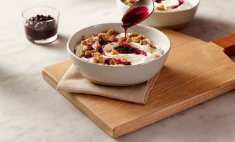 a white bowl filled with granola , yogurt , and fruit is placed on a wooden cutting board , accompanied by a spoon and a cup of wine at SpringHill Suites Atlanta Six Flags