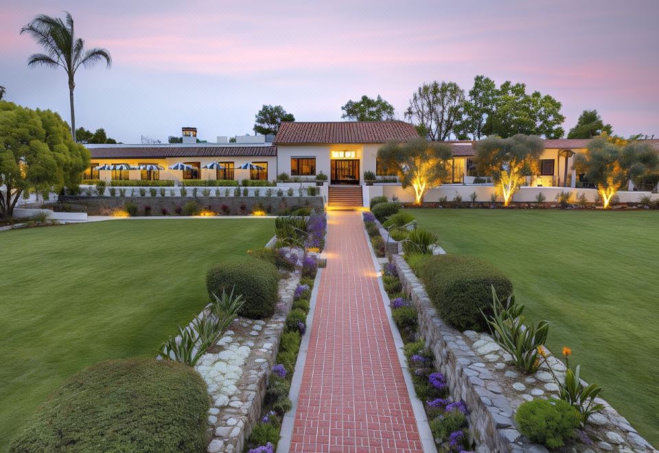 a large , white house with a red brick walkway leading to the entrance , surrounded by lush green grass and trees at The Inn at Rancho Santa Fe