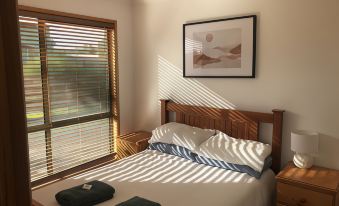 a bedroom with a wooden bed , two pillows , and a window with blinds , bathed in sunlight at Seahorse Coastal Villas