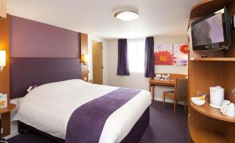a well - lit hotel room with a bed , desk , and window , decorated with purple curtains and flowers at Premier Inn Derby East