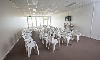an empty conference room with white plastic chairs arranged in rows , facing a podium and a tv at The Island Accommodation