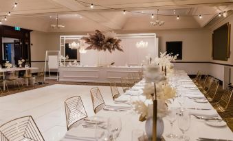 a long dining table is set with white plates , wine glasses , and vases of flowers in front of a bar at Joondalup Resort