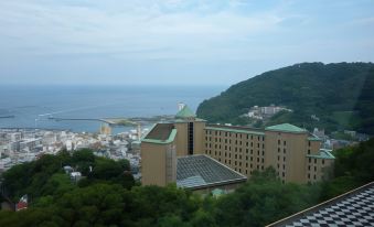 a large building with a green roof is perched on a hill overlooking the ocean at Kamenoi Hotel Atami Annex