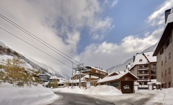 a snow - covered mountain with a ski resort , including a wooden building and ski lifts , under a cloudy sky at Hotel Val de Neu G.L.