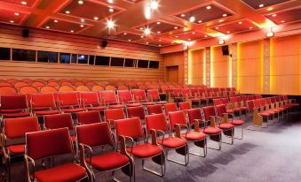 a large conference room with rows of red chairs arranged in a semicircle , and a projector screen mounted on the wall at Novotel Marne la Vallee Noisy le Grand Hotel