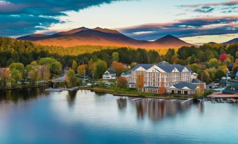 a large , white building is situated on the shore of a lake with mountains in the background at Saranac Waterfront Lodge, Trademark Collection by Wyndham