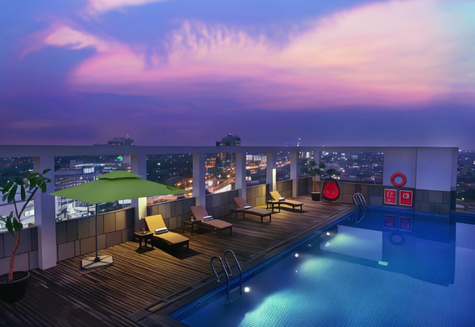 a rooftop pool with lounge chairs and umbrellas , overlooking a cityscape at dusk , as well as the sun sets in the background at ASTON Imperial Bekasi Hotel & Conference Center