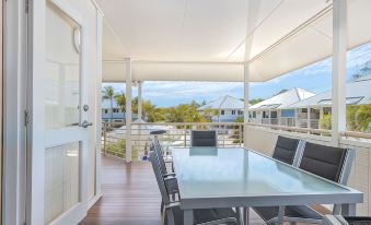 a large glass dining table surrounded by chairs on a wooden deck , with a view of the ocean in the background at Hastings Cove Holiday Apartments