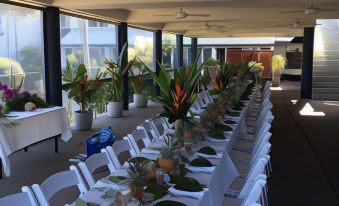 a long dining table is set with white chairs , plates , and a centerpiece of flowers at Rosslyn Bay Resort