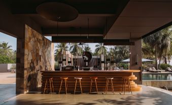 a modern bar with a wooden counter and stools , surrounded by palm trees and a view of the ocean at Karpaha Sands