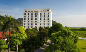 a large white hotel building surrounded by lush green trees and a paved walkway in front of it at Ninh Binh Hidden Charm Hotel & Resort