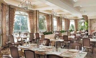 a large dining room with multiple tables and chairs , all set for a meal , is shown with large windows and chandeliers at Swan River Hotel