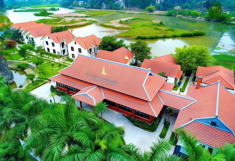 aerial view of a red roofed building surrounded by green grass and trees , with a body of water in the background at Tam Coc La Montagne Resort & Spa