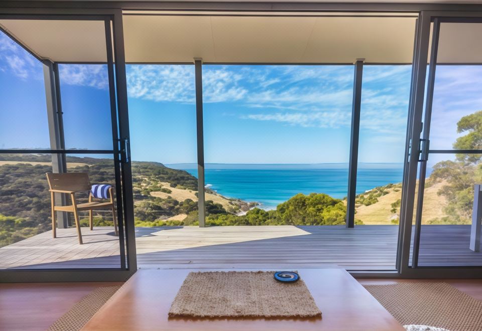 a modern living room with a large window offering a stunning view of the ocean , and a wooden dining table placed on a wooden deck at Sea Dragon Kangaroo Island