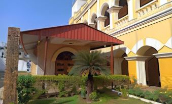 a yellow building with a red awning is surrounded by green grass and palm trees at Hotel Samrat