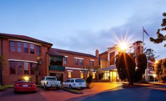 a hotel building with multiple cars parked in front , and the sun setting behind the building at Mercure Maitland Monte Pio