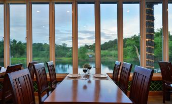 a dining room with a wooden table and chairs set up for a meal , surrounded by large windows overlooking a beautiful lake at Princess River Kwai Hotel
