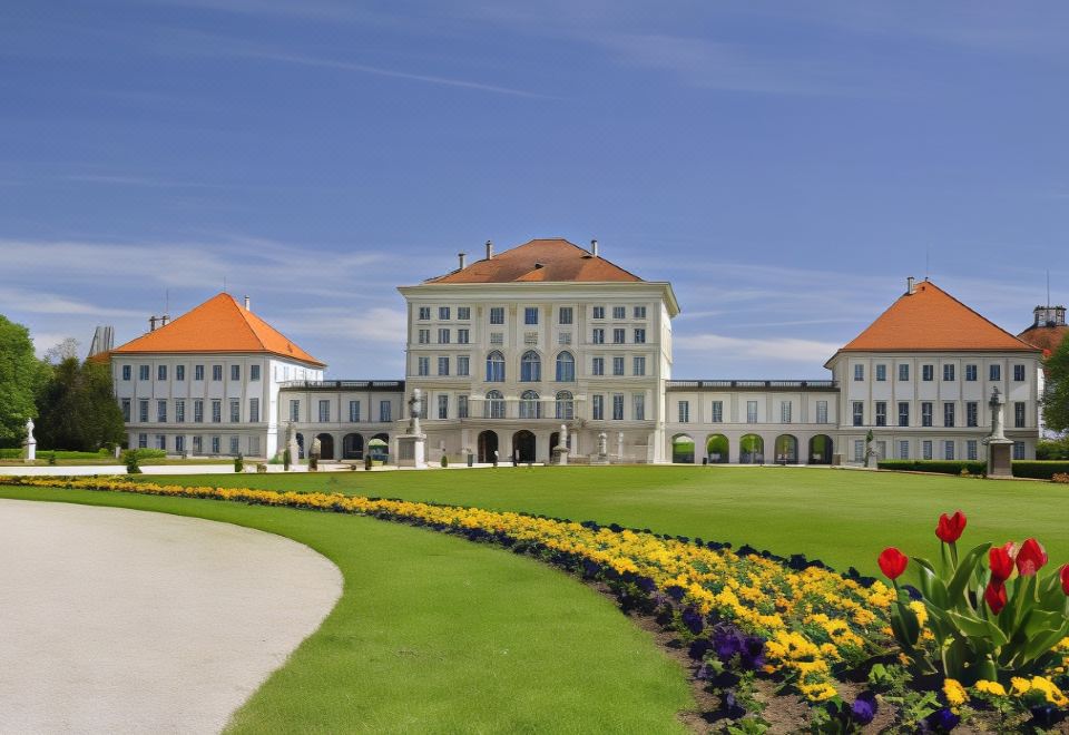 a large , white building with a red roof and orange roof tiles is surrounded by greenery and flower beds at Courtyard Munich Garching