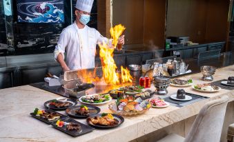 A man is standing in front of a grill, surrounded by food and drinks, as he prepares them at Gems Cube International Hotel