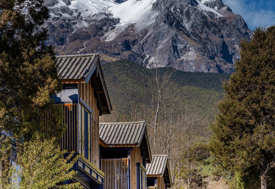 a wooden house surrounded by trees and mountains , with a snow - capped mountain in the background at The Rock Hotel