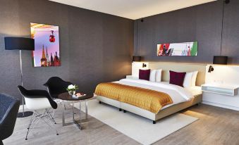 a large bed with a gold blanket is in the middle of a room with a black chair , table , and paintings at Steigenberger Hotel Koln