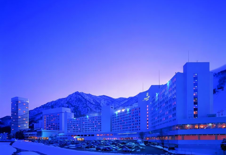 a large hotel building surrounded by snow - covered mountains , with a blue sky above and some lights illuminating the area at Naeba Prince Hotel