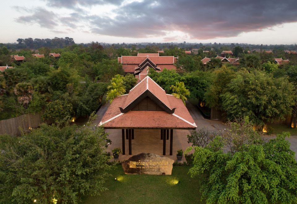 aerial view of a small red roofed house surrounded by trees and grass , with a cloudy sky in the background at Mane Hariharalaya