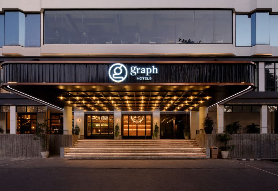 "At night, there is a building with an entrance to an American restaurant and a sign that brightly reads ""Hotel,"" with people coming and going" at Graph Hotels