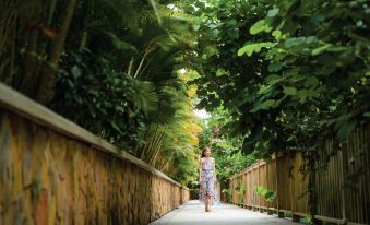 a woman wearing a floral dress is walking down a path lined with palm trees , surrounded by lush greenery at Salinda Resort Phu Quoc - Sparkling Wine Breakfast