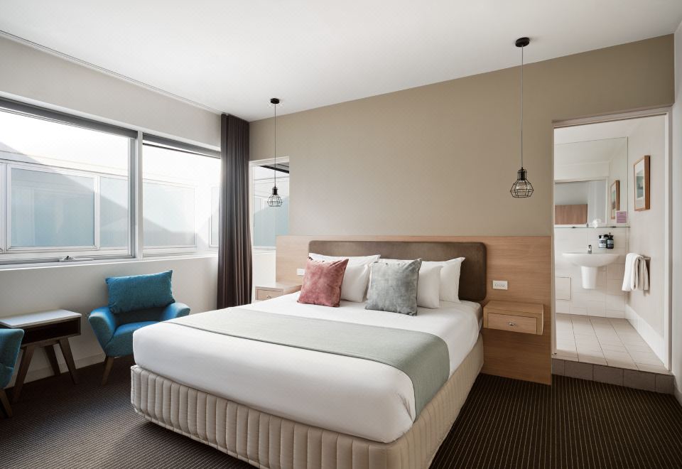 a large bed with a wooden headboard is in a room with a blue chair and windows at Saint Kilda Beach Hotel, an EVT hotel - Formerly Rydges St Kilda