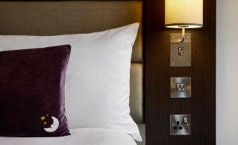 a bed with a white sheet and a purple pillow is next to a wall lamp at Premier Inn Whitehaven