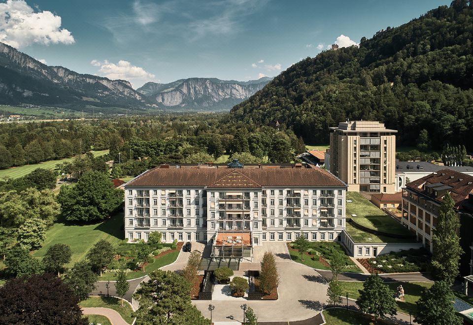 a large , white building with a brown roof and several windows is surrounded by greenery and mountains at Grand Resort Bad Ragaz