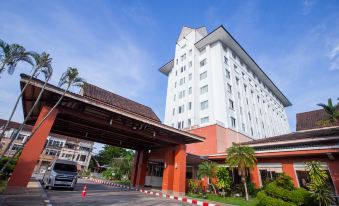 a large white building with a red roof , situated in a parking lot surrounded by trees at The Imperial Narathiwat Hotel