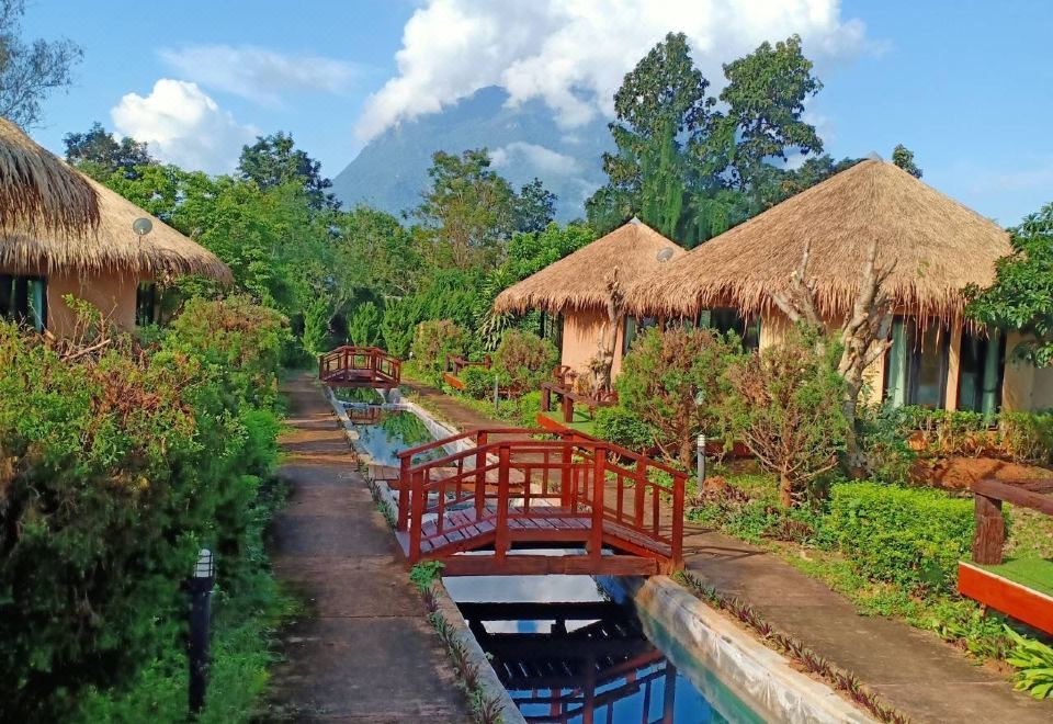 a picturesque scene of a small wooden bridge spanning a body of water , surrounded by lush greenery and thatched - roof huts at Aurora Resort Chiangdao