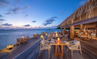 a beachfront restaurant with wooden tables and chairs , straw umbrellas , and a view of the ocean at sunset at Anantara Kihavah Maldives Villas
