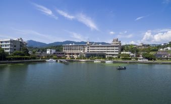 a large building is situated on the shore of a lake with mountains in the background at Biwako Ryokusuitei