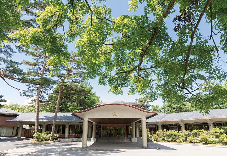 a large building with a covered entrance and trees in the background , under a clear blue sky at Karuizawa Prince Hotel East
