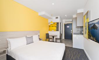 a modern hotel room with yellow walls , white bed , and kitchenette , along with a dining area and small kitchenette at ValueSuites Green Square