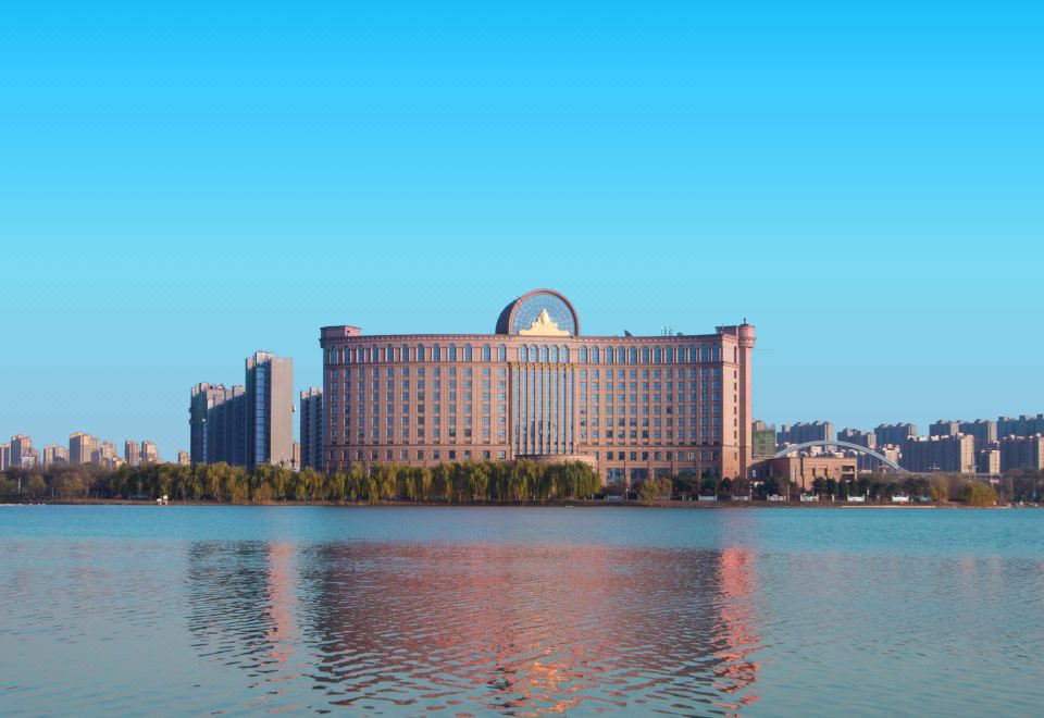 A large building is reflected in water with the city behind it, as seen from across at Grand Metropark Guofeng Hotel, Tangshan