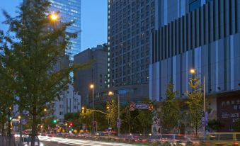 At night, a city street is lined with tall buildings and pedestrians, while an empty sidewalk can be seen nearby at Swissotel Grand Shanghai