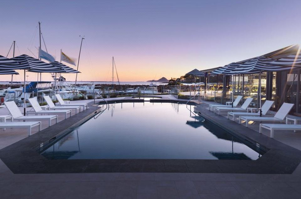 a large outdoor pool surrounded by lounge chairs and umbrellas , with the sun setting in the background at Anchorage Port Stephens