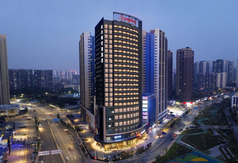 A large building stands in the center, surrounded by other buildings, in a night time lapse photo at Hampton by Hilton Shenzhen North Station