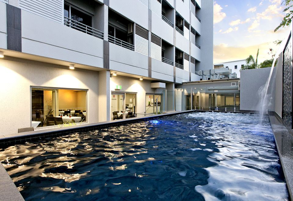 a modern hotel with a swimming pool , surrounded by white buildings and illuminated by lights at Argus Hotel Darwin