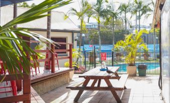 a wooden bench and table on a patio with palm trees in the background , overlooking a pool at Brisbane Backpackers Resort