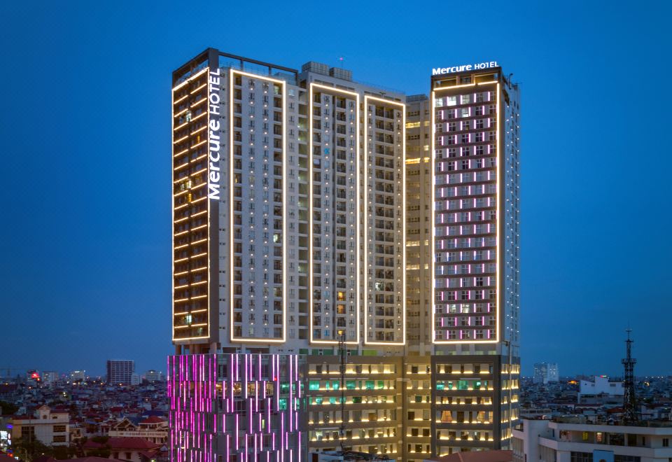 a tall building with a pink and blue light show on its side , surrounded by other buildings in a city at Mercure Hai Phong