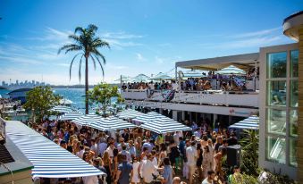 a large crowd of people gathered on a beach , some standing and others sitting under striped umbrellas at Watsons Bay Boutique Hotel
